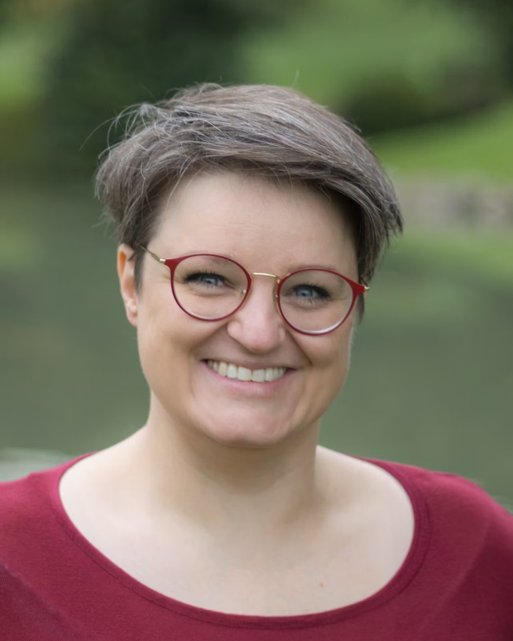 Kai Buder, Pädagogin, lächelt in die Kamera, kurzes Haar, rote Brille, roter Pullover, im Hintergrund unscharfes Grün