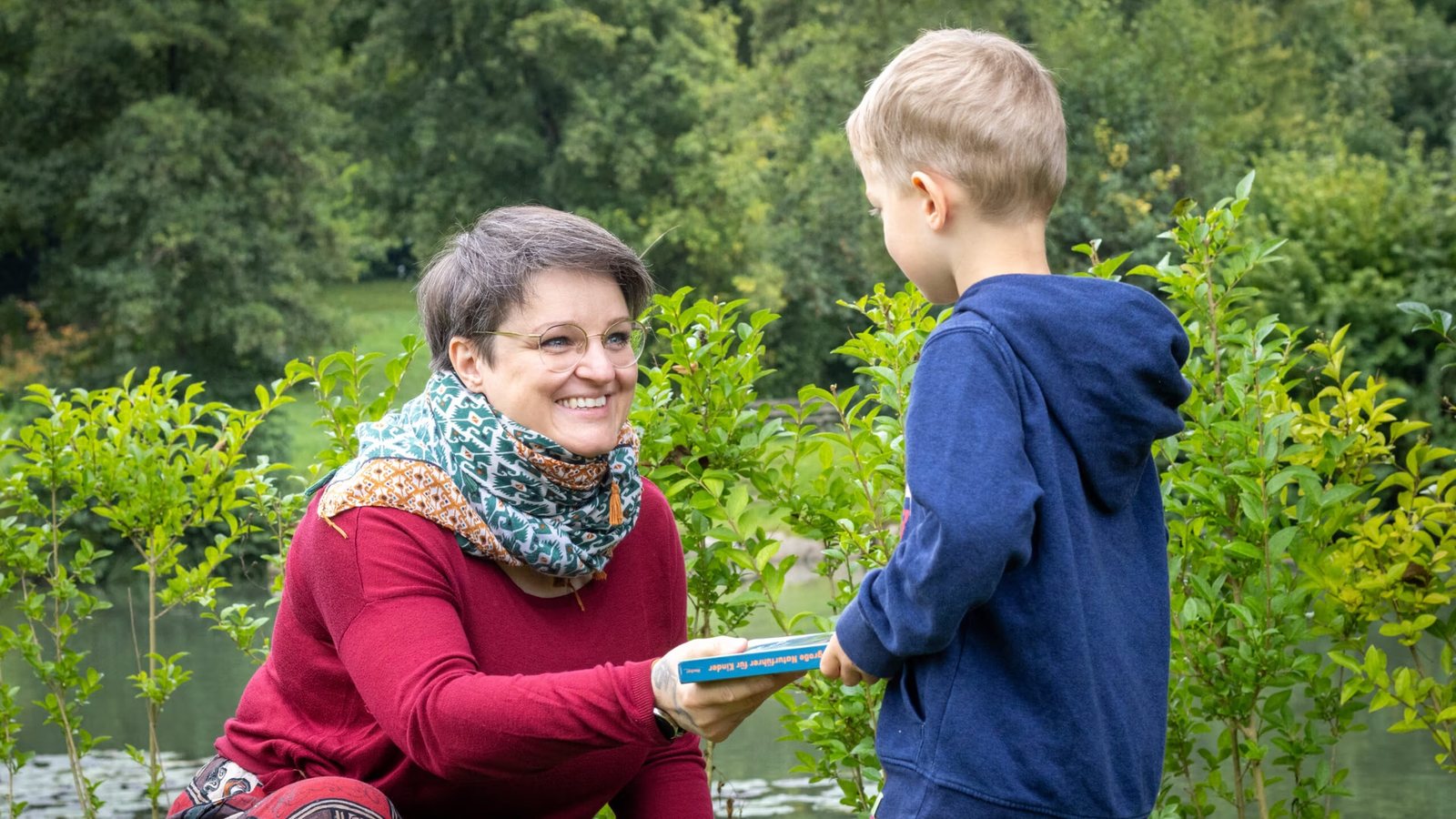 Kai Buder kniet lachend vor einem Kind und hält mit ihm ein Buch, im Hintergrund grüne Sträucher und Wasser