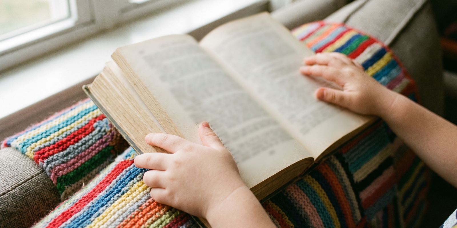 Kinderhände halten ein dickes Buch ohne Bilder auf einer bunten Strickdecke